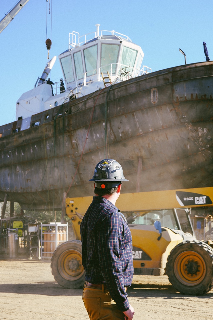 Thompson Marine Employee working in marine industry wearing hardhat that reads Thompson Strong, standing in front of CAT equipment and ship being repaired in shipyard.